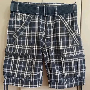  rutiga cargoshorts  - Rutiga cargoshorts i marinblå och vitt med stora fickor på sidorna och bak. Shortsen har ett medföljande svart bälte och snörning nertill. Perfekta för dig som gillar en avslappnad och cool stil. Materialet känns som bomull och passformen är loose.