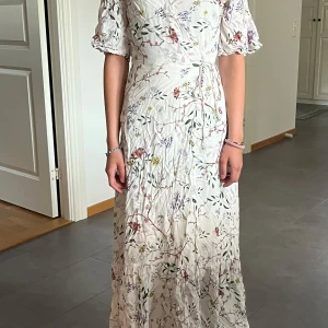 Vit blommig långklänning H&M - Superfin långklänning från H&M i vitt med blommigt mönster i grönt, lila och rosa. Klänningen har korta puffärmar, omlottknytning i midjan och volang nertill. Perfekt för sommarens alla tillfällen!