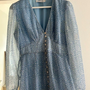 Blå leopardmönstrad långklänning Adoore - Supersnygg långklänning från Adoore i ljusblått med vitt leopardmönster. Klänningen har v-ringning, knappar framtill och långa transparenta ärmar. Perfekt för dig som vill sticka ut med ett coolt mönster och en feminin siluett.