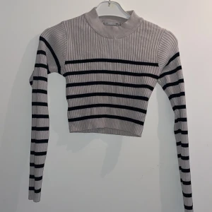 Randig croppad ribbad tröja - Säljer en croppad ribbad tröja med svarta och gråa horisontella ränder. Tröjan har lång ärm och hög hals, perfekt för dig som gillar en trendig och stilren look. Materialet är stretchigt och sitter snyggt på kroppen.