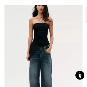 Svart asymmetrisk topp från Zara - Supersnygg svart topp från Zara med asymmetrisk skärning och draperad framsida. Toppen är axelbandslös och har en cool, modern look som verkligen sticker ut. Perfekt för dig som vill ha något unikt och trendigt. Helt oanvänd