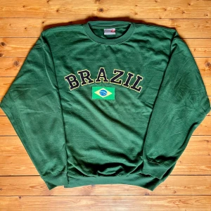 Grön Brazil sweatshirt Nick's World XXL - Grön sweatshirt från Nick's World i storlek XXL med stort 'BRAZIL'-tryck och brasiliansk flagga på bröstet. Klassisk rund halsringning och långa ärmar. Perfekt för dig som vill sticka ut med sportig vibe och visa kärlek till Brasilien.
