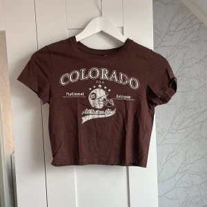 Brun croppad Colorado t-shirt H&M - Snygg brun croppad t-shirt från H&M Divided med tryck 'Colorado National League' och amerikansk fotbollshjälm framtill. T-shirten har korta ärmar, rund hals och är gjord i mjuk bomull. Perfekt för en sportig och avslappnad stil.