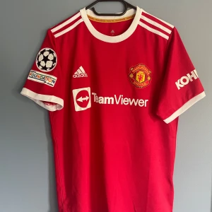Manchester United Ronaldo 7 Adidas tröja - Röd Manchester United matchtröja med vita detaljer, officiella tryck för Ronaldo 7 på ryggen och Champions League-märke på ärmen. Adidas-logga och TeamViewer sponsor framtill. Tillverkad i lätt och ventilerande polyester, klassisk passform med korta ärmar och rund hals.