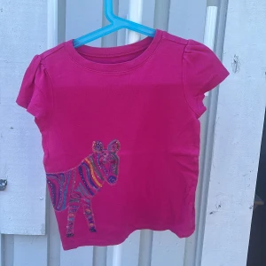 Rosa t-shirt med zebra från Gap Kids - Snygg rosa t-shirt från Gap Kids med korta ärmar och färgglad zebraprint med paljettdetaljer på både fram- och baksidan. T-shirten är i mjuk bomull och har en rund halsringning. Perfekt för dig som gillar färg och coola djurmotiv!