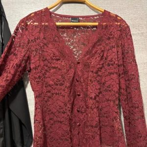 Vinröd spetsblus från Gina Tricot - Supersnygg vinröd spetsblus från Gina Tricot med v-ringning och klädda knappar framtill. Blusen har långa ärmar med lätt utsvängda ärmslut och är helt i genombruten spets med blommigt mönster. Perfekt för dig som vill sticka ut lite extra!