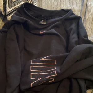 Svart Nike sweatshirt med rosa logga - Svart sweatshirt från Nike med rosa swoosh på bröstet och stort Nike-tryck i rosa . Perfekt för en sportig och avslappnad stil. Storlek L 