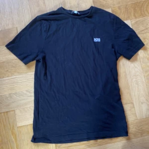 Svart t-shirt från Hugo Boss - Svart t-shirt från Hugo Boss med diskret logga på bröstet. Klassisk rund halsringning och korta ärmar. Tillverkad i mjuk bomull som känns skön mot huden. Perfekt för en clean och stilren look.