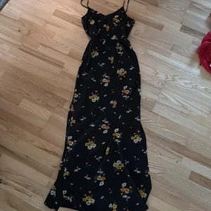 Svart blommig långklänning - Svart långklänning med smala axelband och blommigt mönster i gult och vitt. Klänningen har en feminin siluett och är perfekt för varma dagar. Lätt och luftig känsla, passar dig som gillar romantisk stil. med slits längs benet