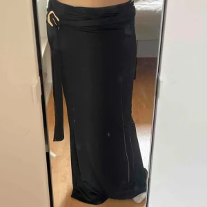 Svart långkjol med knyt i midjan - Säljer en svart långkjol med snygg knytning och bältedetalj i midjan. Kjolen har en rak och elegant passform som sitter skönt på kroppen. Perfekt för dig som gillar stilrena och enkla plagg med en twist.