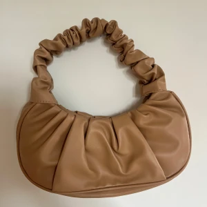 Beige axelväska med rynkad rem - Snygg beige axelväska i syntetmaterial med cool rynkad rem och veckad design framtill. Väskan har en mjuk, rundad form och är perfekt för att lyfta din outfit med en trendig touch.