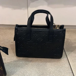 Svart handväska i skinn - Snygg svart handväska  i präglat skinn . Väskan har två handtag, guldfärgade metalldetaljer och dragkedja upptill. Perfekt storlek för det viktigaste och en clean, lyxig look.