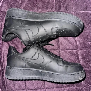 Svarta Nike Air Force 1 sneakers - Klassiska helsvarta Nike Air Force 1 sneakers i skinn med perforerad tå och svart sula. Snygg och clean look med diskret Nike-logga på sidan och metallbricka med 'AF1' på snörningen. Perfekta för dig som gillar streetstyle och tidlös design.