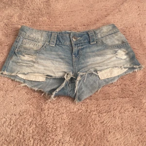 Ljusblå slitna jeansshorts Gina Tricot +14 - Snygga ljusblå jeansshorts från Gina Tricot med slitna detaljer och fransig kant. Klassisk femficksmodell med bälteshällor och knappstängning. Perfekta för varma dagar när du vill ha en avslappnad och trendig look.