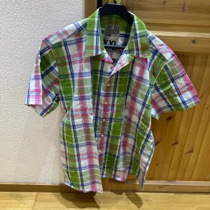 Färgglad rutig skjorta från JMY DEE - Kortärmad skjorta från JMY DEE i bomull med rutigt mönster i grönt, rosa, blått och vitt. Skjortan har klassisk krage och knäppning framtill. 