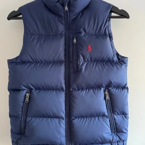 Blå dunväst från Polo Ralph Lauren storlek 134/140 - Snygg marinblå dunväst från Polo Ralph Lauren med klassisk röd logga på bröstet. Västen har tre dragkedjeförsedda fickor och hög krage. Quiltad design och stilren look.
