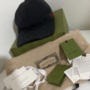 Gucci keps - Snygg svart keps från Gucci med diskret GG-monogram över hela tyget och klassiska gröna och röda ränder på sidan. Kepsen har böjd skärm och justerbar rem bak. Kommer med originalbox, dustbag och tags. Perfekt för dig som vill ha en exklusiv streetstyle-look.