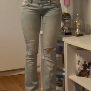 Ljusblå bootcut jeans med slitning - Säljer ett par ljusblå bootcut jeans med låg midja och snygg slitning på ena knät. Jeansen har olika detaljer på sig. 
