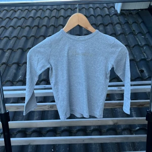 Grå långärmad tröja från Benetton - Säljer en stilren grå långärmad tröja från Benetton i mjuk ekologisk bomull. Tröjan har en enkel design med rund halsringning och diskret broderad text framtill. Perfekt för dig som gillar basic och bekvämt.
