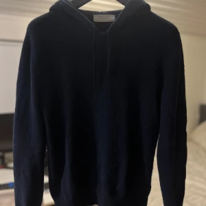 Mörkblå hoodie i kashmir - Mysig mörkblå hoodie från sayless i lyxig kashmir. Tröjan har huva med snörning och långa ärmar, perfekt för dig som gillar en clean och stilren look. Passformen är normal och materialet är supermjukt och skönt mot huden. Ett litet hål under armen se på bild 3.