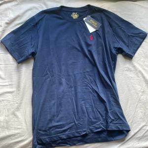 Marinblå t-shirt Polo Ralph Lauren - Snygg marinblå t-shirt från Polo Ralph Lauren i bomull med röd broderad logga på bröstet. Modellen är slim fit och har klassisk rund hals och korta ärmar. Helt oanvänd.