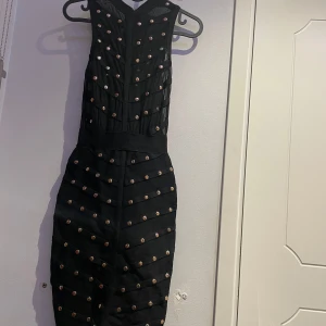 Svart kort klänning med nitar - Snygg svart kort klänning med tight passform och ärmlös design. Klänningen har coola nitar i guld och silver som dekoration över hela framsidan och ryggen. Klänningen har dragkedja bak. Perfekt för dig som gillar edgy stil. Använd 2 ggr. Kommer inge ihåg var den är köpt men den köptes flr ca 800. Pris kan diskuteras 