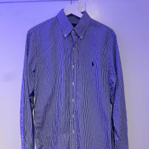 Randig blå skjorta från Ralph Lauren - Snygg slim fit skjorta från Ralph Lauren i blått och vitt med ränder. Skjortan har lång ärm, knappar framtill och den ikoniska broderade Polo-loggan på bröstet. Perfekt krage och stilren design som ger en fräsch look.