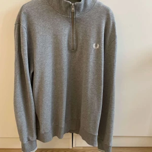 Grå half zip Fred Perry  - Snygg grå sweatshirt från Fred Perry i storlek XL men passar L. Tröjan har half zip framtill, hög krage och långärmade ribbade muddar. Broderad Fred Perry-logga på bröstet. Perfekt för chill dagar och enkel att matcha med jeans eller joggers. Haft den i ca 3-4 månader men inte kommit till mycket användning, skick 9/10. 