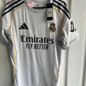 Real Madrid Rodrygo 11 matchtröja - Snygg vit Real Madrid fotbollströja från Adidas med Rodrygo och nummer 11 på ryggen. Tröjan har svarta detaljer, gula linjer och klubbmärke på bröstet. Kortärmad med Champions League-märke på ärmen och tillverkad i lätt funktionsmaterial.