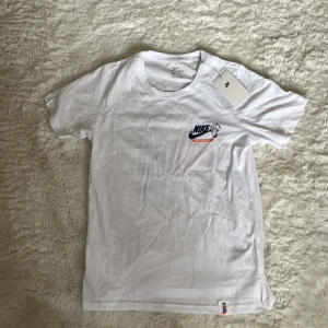 Vit Nike Dri-Fit t-shirt med tryck - Vit Nike Dri-Fit t-shirt med coolt tryck på ryggen och liten logga framtill. T-shirten har rund hals, korta ärmar och är gjord i ett lätt och sportigt material. Perfekt för dig som gillar streetwear och sneakers.