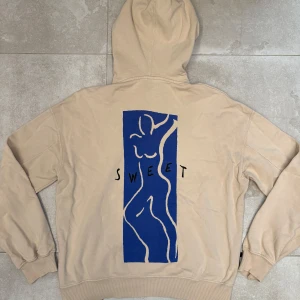 Beige hoodie från Sweet SKBTS  - Beige hoodie från Sweet SKBTS med huva och snörning, samt känguruficka framtill. På ryggen finns ett stort blått konstinspirerat tryck med en stiliserad kropp och texten SWEET. Relaxed oversize fit. (passar S/M)