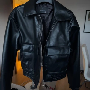 Svart skinnjacka från Stradivarius - Snygg svart skinnjacka från Stradivarius i klassisk modell med stora fickor framtill och dragkedja. Jackan har bred krage, elastisk nederkant och raka ärmar. Perfekt för dig som vill ha en stilren och edgy look.