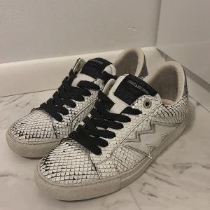 Zadig & Voltaire sneakers i ormskinnsmönster - Snygga sneakers från Zadig & Voltaire med vit och silverfärgad ovandel i ormskinnsmönstrat skinn. Skorna har svarta snören, rund tå och en cool blixtdetalj på sidan. Perfekta för dig som vill sticka ut med en unik och trendig look.