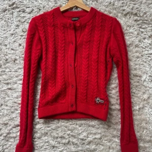 Röd stickad kofta från Napapijri - Riktigt snygg röd kofta från Napapijri med kabelstickat mönster och knappar framtill. Långärmad och med norsk flagga broderad nertill. Perfekt för dig som gillar klassisk stil med en twist. Materialet är mjukt och stickat, känns både varmt och trendigt.