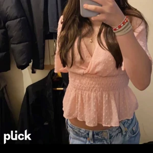 Ljusrosa blus med volang och v-ringning - Supersöt ljusrosa blus med v-ringning och omlott framtill. Blusen har korta ärmar, smockad midja och volang nertill. Tunt, prickigt tyg som ger en romantisk vibe. Perfekt att matcha med jeans eller kjol.