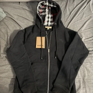 Svart hoodie från Burberry med dragkedja - Svart hoodie från Burberry med klassiskt rutigt foder i huvan, silverfärgad dragkedja och snörning. Broderad logga på bröstet och ribbade muddar. Perfekt för dig som vill ha en stilren och lyxig streetwear-look.