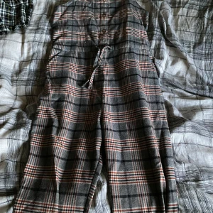 Rutig jumpsuit från Next med snörning - Säljer en rutig jumpsuit från Next i grått, svart och rött. Modellen har snörning i midjan och elastisk rygg för skön passform. Materialet är mjukt med polyester, viskos och lite elastan. Perfekt för dig som gillar chill och snygg stil.