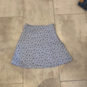 Ljusblå blommig kortkjol med dragkedja - Supersöt ljusblå kortkjol med små mörkblå blommor och diskreta vita detaljer. Kjolen har en smickrande A-linjeform och en praktisk dragkedja i sidan. Perfekt för dig som gillar en feminin och fräsch stil under varmare dagar.