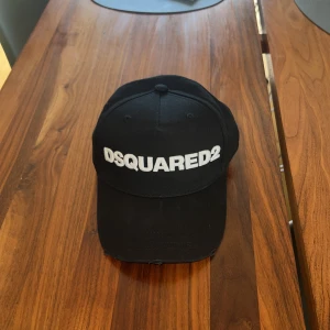 Svart keps från Dsquared2 - Snygg svart keps från Dsquared2 med vit broderad logga framtill och texten 'Dean & Dan Caten' baktill. Justerbart spänne för perfekt passform. Klassisk böjd skärm och stilren design som passar till allt. Aldrig använd. 