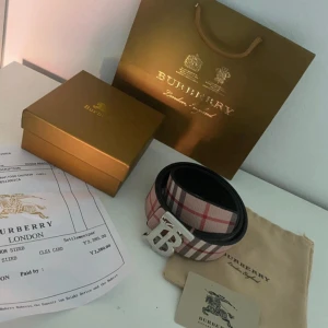Rutig Burberry bälte med silverdetalj - Snyggt Burberry bälte med klassiskt rutigt mönster i beige, rött och svart. Bältet har en stor silverfärgad spänne med Burberrys ikoniska TB-logo. Utsidan är i canvas med rutor och insidan i svart skinn. Perfekt accessoar för att lyfta din outfit.