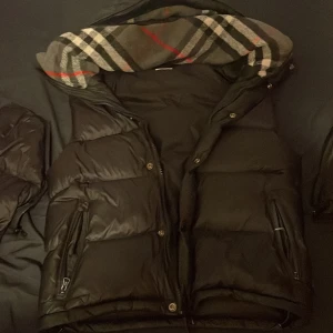 Svart dunväst från Burberry - Säljer en svart dunväst från Burberry med klassiskt rutigt foder i huvan. Västen har dragkedja, knappar och två fickor med dragkedja. Stort Burberry-märke på ena sidan och ikoniskt rutmönster i grått, vitt och rött på insidan av huvan. Väldigt ny har användt några månader bara 
