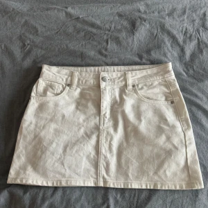 Vit jeanskjol med fickor - Säljer en vit jeanskjol med klassisk femficksdesign och knappstängning framtill. Kjolen är kort och har en rak passform som ger en clean och fräsch look. Perfekt att matcha med sneakers eller boots för en chill stil.