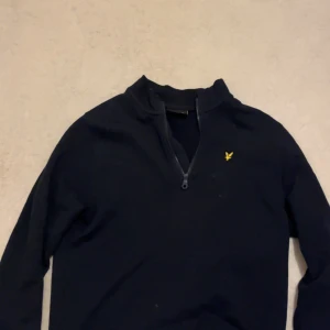 Marinblå half zip tröja Lyle & Scott - Snygg marinblå långärmad tröja från Lyle & Scott med half zip och klassisk gul logga på bröstet. Tröjan har en stilren design och är gjord i mjuk bomull, perfekt för dig som gillar enkel och clean stil. Passar dig som vill ha något bekvämt och coolt.