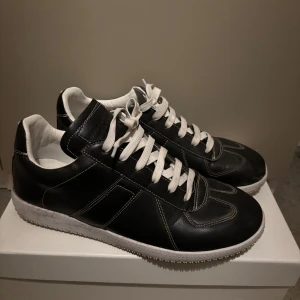 Maison Margiela svarta sneakers - Sällsynta Maison Margiela sneakers i svart skinn med vita kontrastsömmar och vita snören. Klassisk rund tå och platt sula, insida i vitt och detaljerad yttersula. Snygg minimalistisk design som passar till streetwear och modeintresserade.