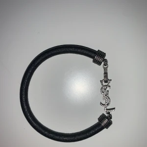 Svart YSL armband med silverdetaljer - Snyggt svart armband från YSL med grov läderrem och coola silverdetaljer. Armbandet har ett dekorativt YSL-logo-hänge i silver med små nitar och textur. Perfekt accessoar för dig som gillar lyxiga märken och stilrena smycken.