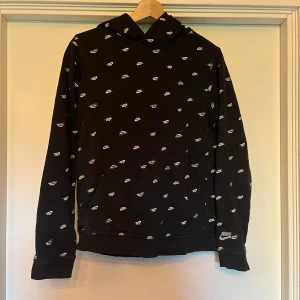 Svart Nike hoodie med loggor - Svart hoodie från Nike i storlek 158-170, täckt med små vita Nike-loggor över hela tröjan. Klassisk känguruficka framtill och huva. Perfekt för dig som gillar sportig och street-inspirerad stil.