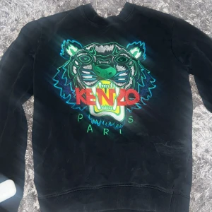 Svart Kenzo sweatshirt med tiger - Svart sweatshirt från Kenzo med ikoniskt broderat tigerhuvud i blått, grönt och vitt samt röd Kenzo-logga på bröstet. Tröjan har rund hals, långa ärmar och ribbade muddar. Perfekt statement-plagg för dig som gillar streetstyle.