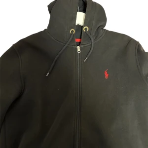 Svart hoodie  - Svart hoodie från Polo Ralph Lauren med dragkedja och huva. Klassisk röd broderad logga på bröstet och snörning i huvan. Mjuk insida och ribbade muddar. Perfekt för en chill och stilren look.
