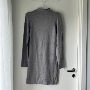 Grå stickad klänning French Connection - En stilren grå stickad klänning från French Connection i storlek M. Klänningen har lång ärm, rak passform och en enkel hög krage. Perfekt för dig som gillar minimalistisk och clean stil.