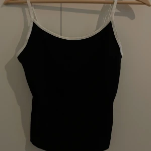 Svart linnetopp med vita kanter - En stilren svart linnetopp med tunna axelband och kontrasterande vita kanter. Toppen har en enkel och rak passform som gör den lätt att matcha med olika outfits. Perfekt för varma dagar eller som lager under en skjorta.